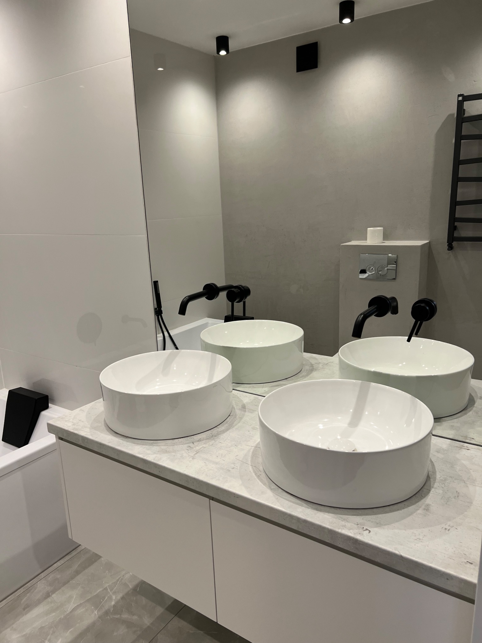 Nowoczesna łazienka z dwoma okrągłymi, białymi umywalkami nablatowymi z czarnymi bateriami, szara ściana z toaletą podtynkową, białe płytki na ścianie obok wanny i czarny grzejnik drabinkowy.