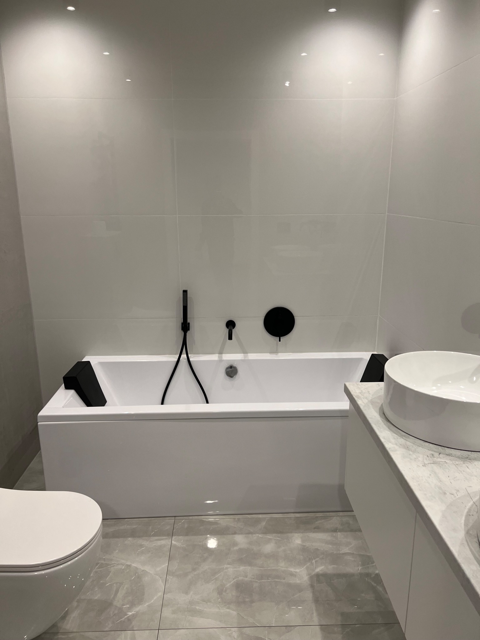 Minimalistyczna łazienka w odcieniach bieli i szarości z czarnymi akcentami: biała wanna z czarną armaturą, umywalka nablatowa na blacie imitującym marmur i podwieszana toaleta.