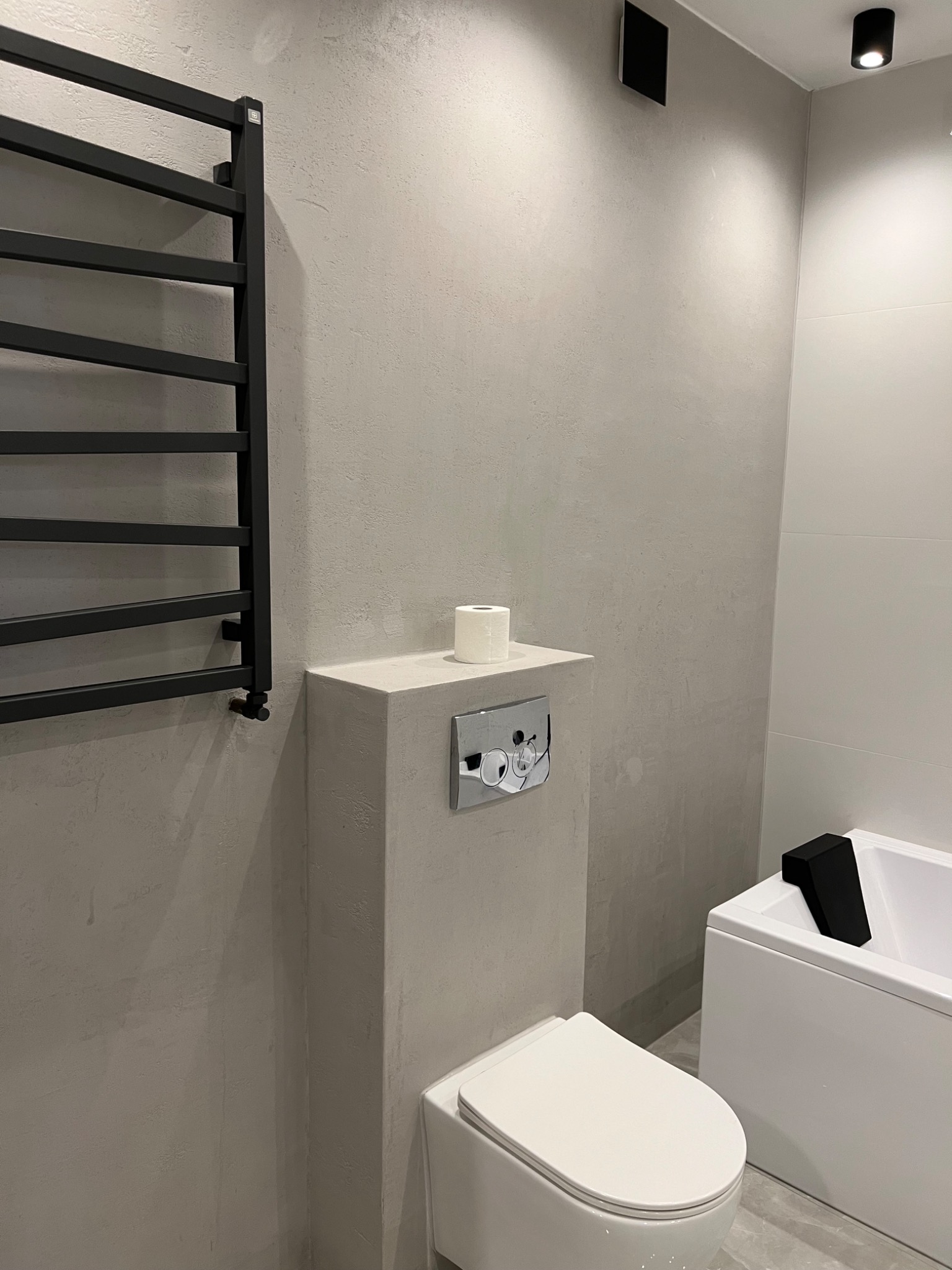 Nowoczesna łazienka w odcieniach szarości z widoczną teksturą ścian, minimalistyczna toaleta podwieszana z chromowanym przyciskiem spłukującym, czarny grzejnik drabinkowy i fragment białej wanny...