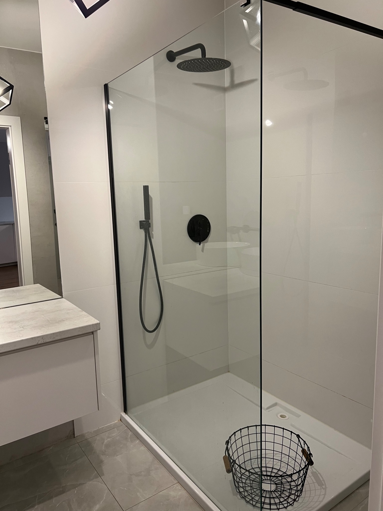 Nowoczesna kabina prysznicowa z czarnymi elementami armatury, szklaną ścianą i koszem na akcesoria w rogu, białe płytki na ścianach i szara marmurowa podłoga.