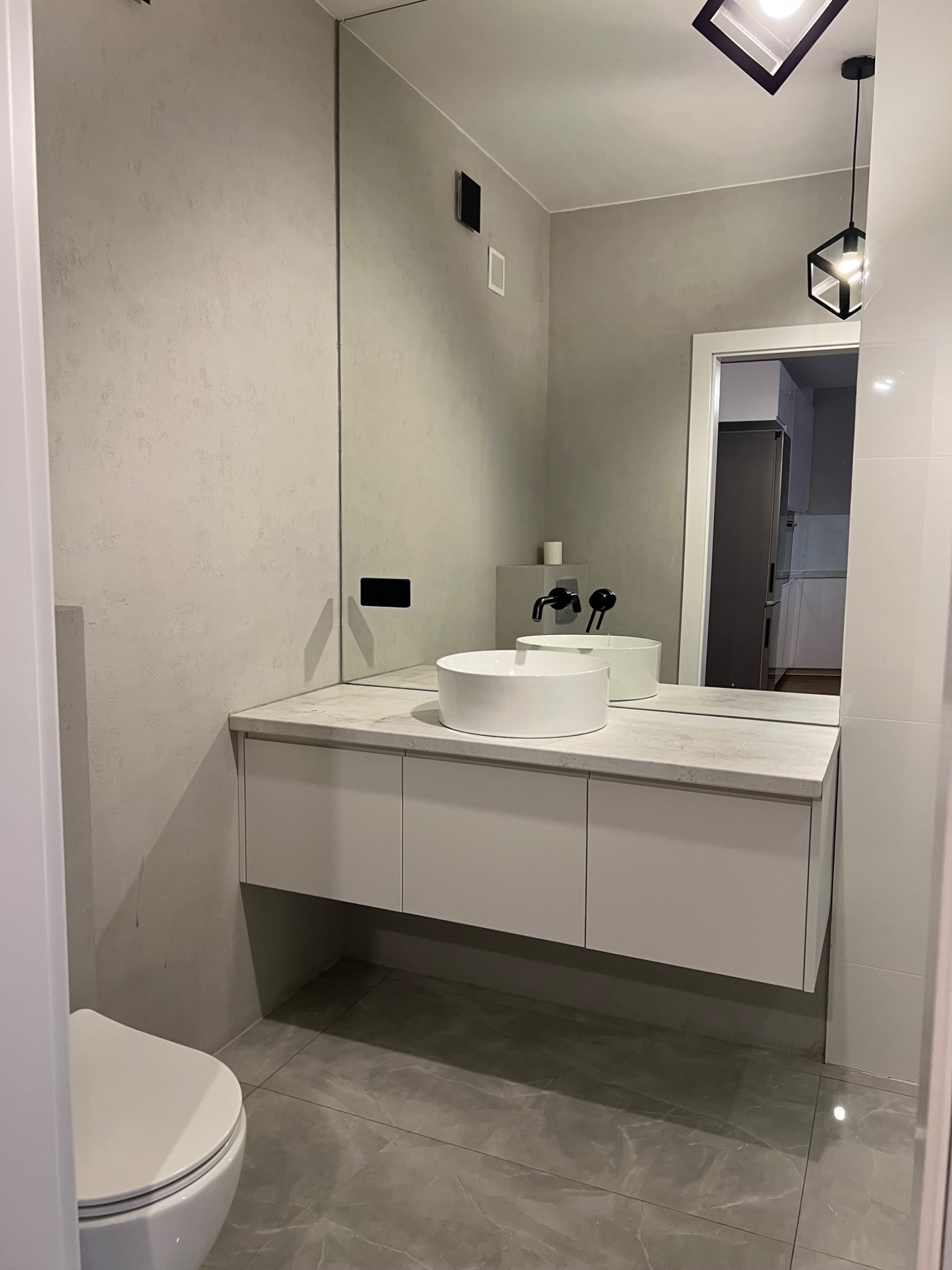 Nowoczesna łazienka z umywalką nablatową, szarą ścianą z efektem betonu, dużym lustrem i geometryczną lampą wiszącą, widoczny fragment toalety i płytek podłogowych.