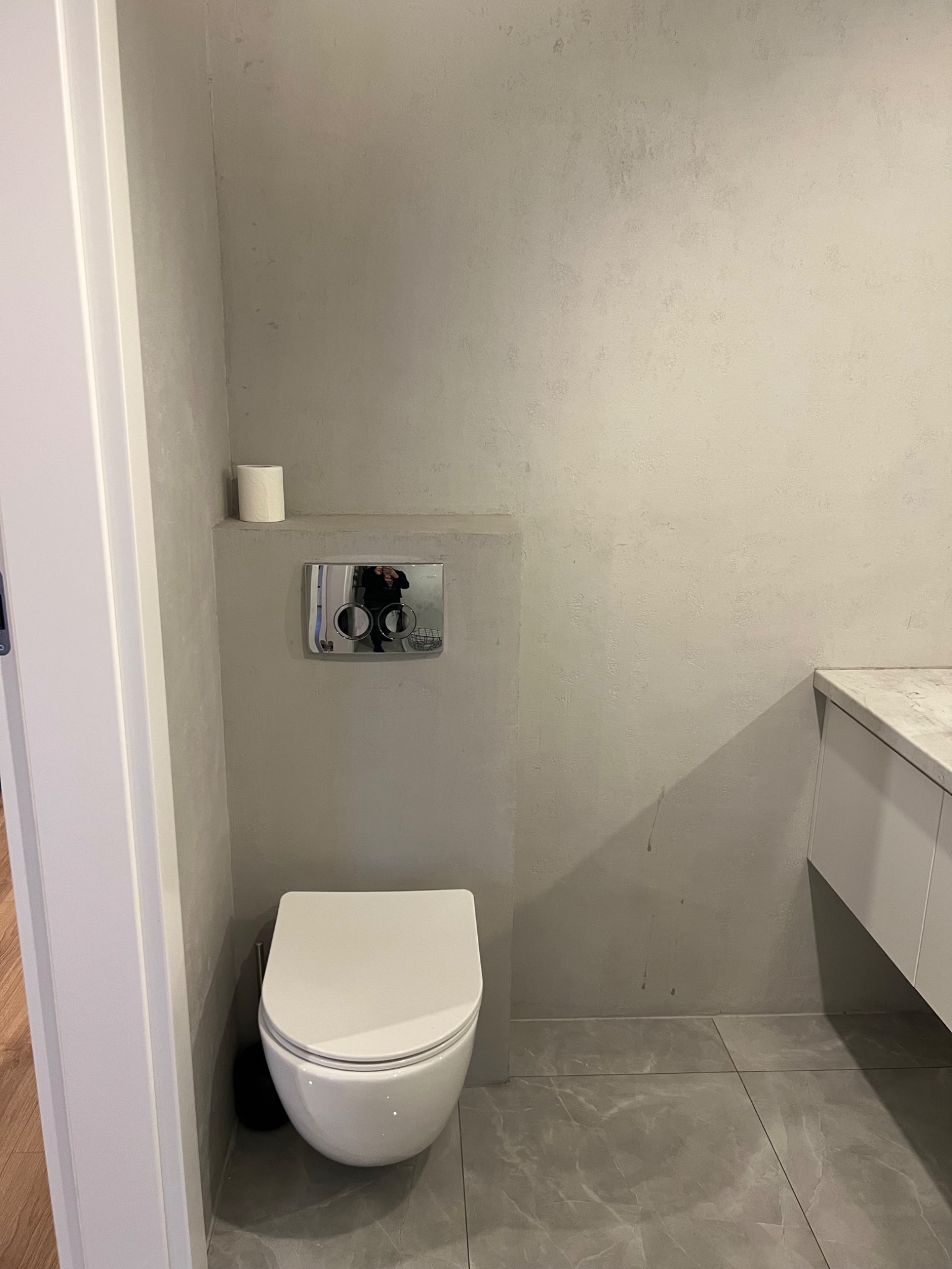 Nowoczesna toaleta wisząca w minimalistycznej łazience z szarą, teksturowaną ścianą, marmurową podłogą i białymi meblami łazienkowymi.