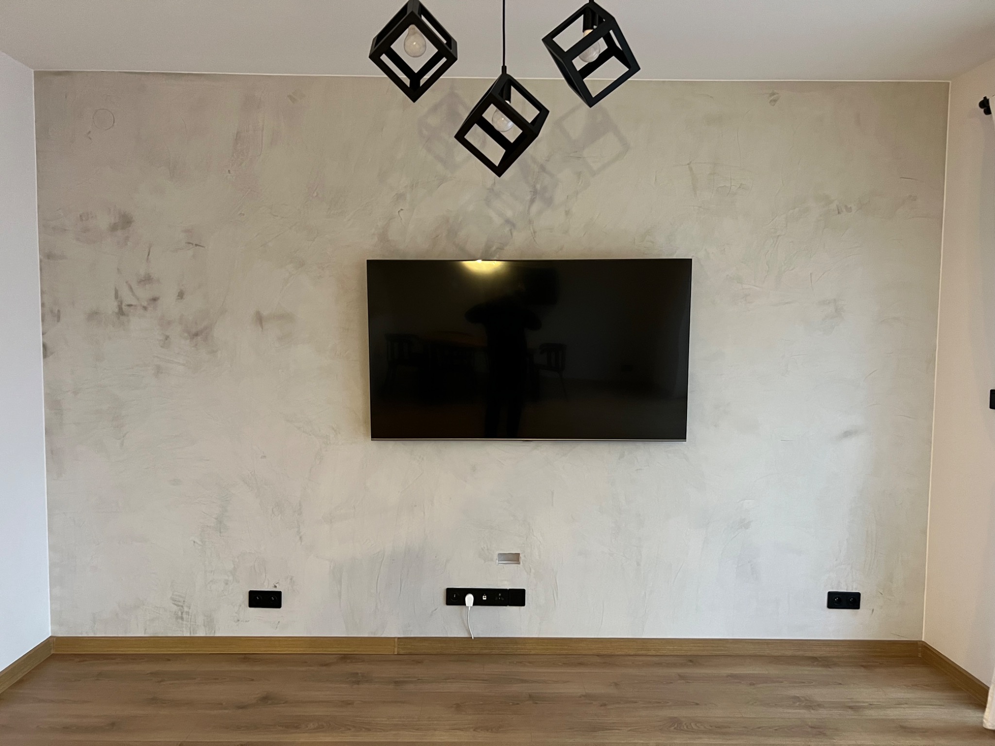 Ściana w salonie z efektem dekoracyjnym, podwieszony telewizor, nowoczesne lampy w kształcie czarnych sześcianów, widoczne gniazdka elektryczne przy podłodze.