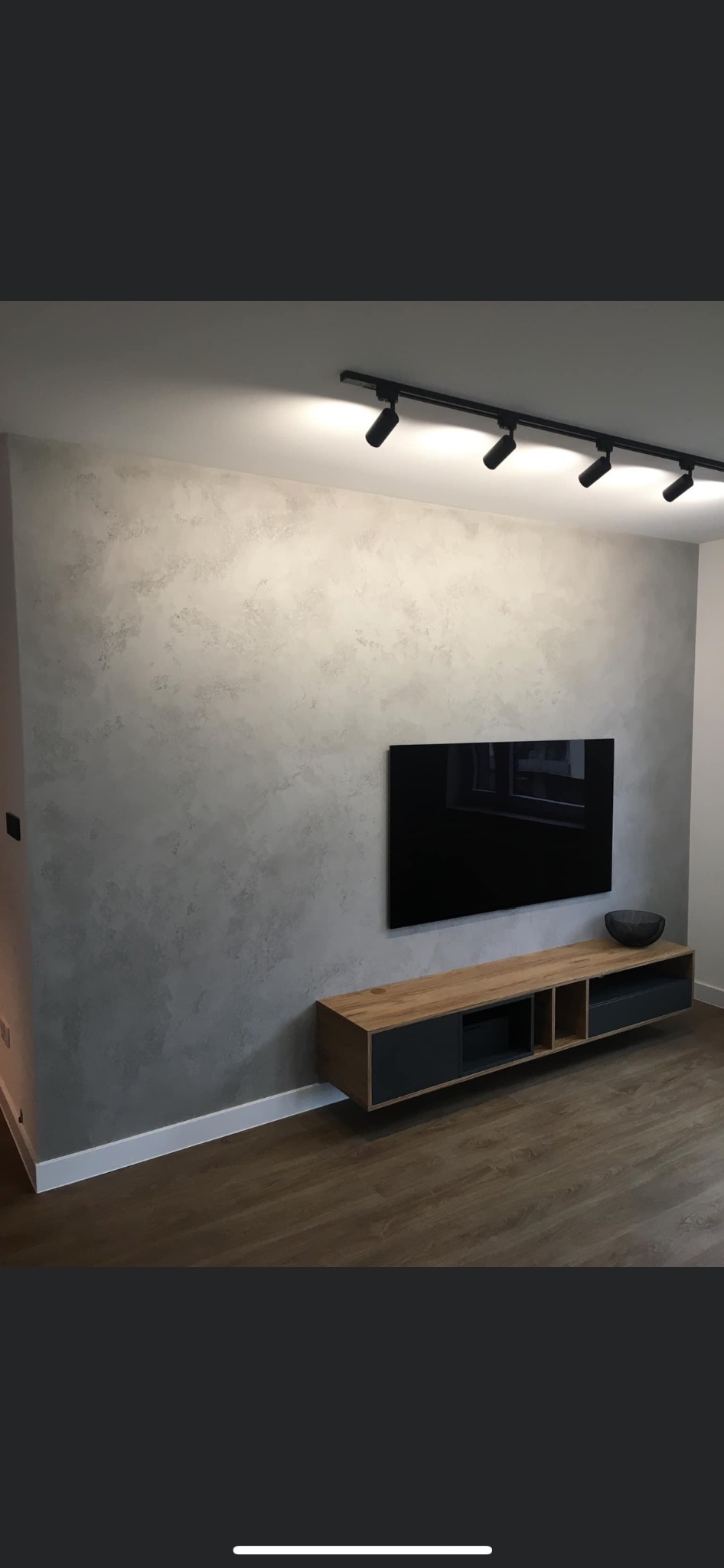 Ściana wykończona dekoracyjną strukturą w odcieniach szarości, oświetlona listwą z reflektorami, z zawieszonym telewizorem i szafką RTV w stylu nowoczesnym.