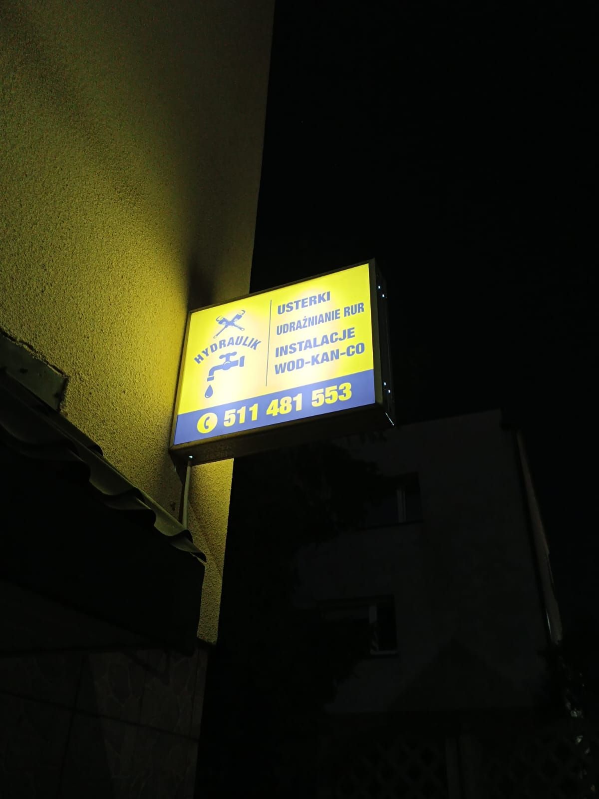 Oświetlony szyld reklamowy firmy hydraulicznej z numerem telefonu, zamontowany na elewacji budynku w nocnym ujęciu.