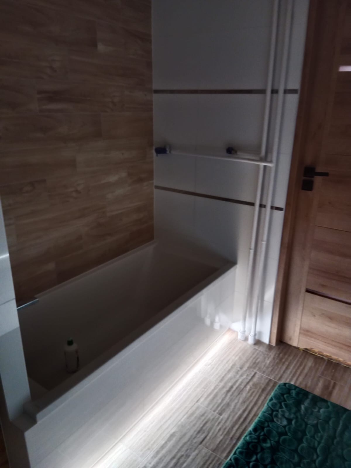 Wykończona łazienka z wanną podświetloną od dołu, drewnopodobne płytki na ścianach i podłodze, widoczny fragment zielonego dywanika i butelka z płynem do kąpieli.