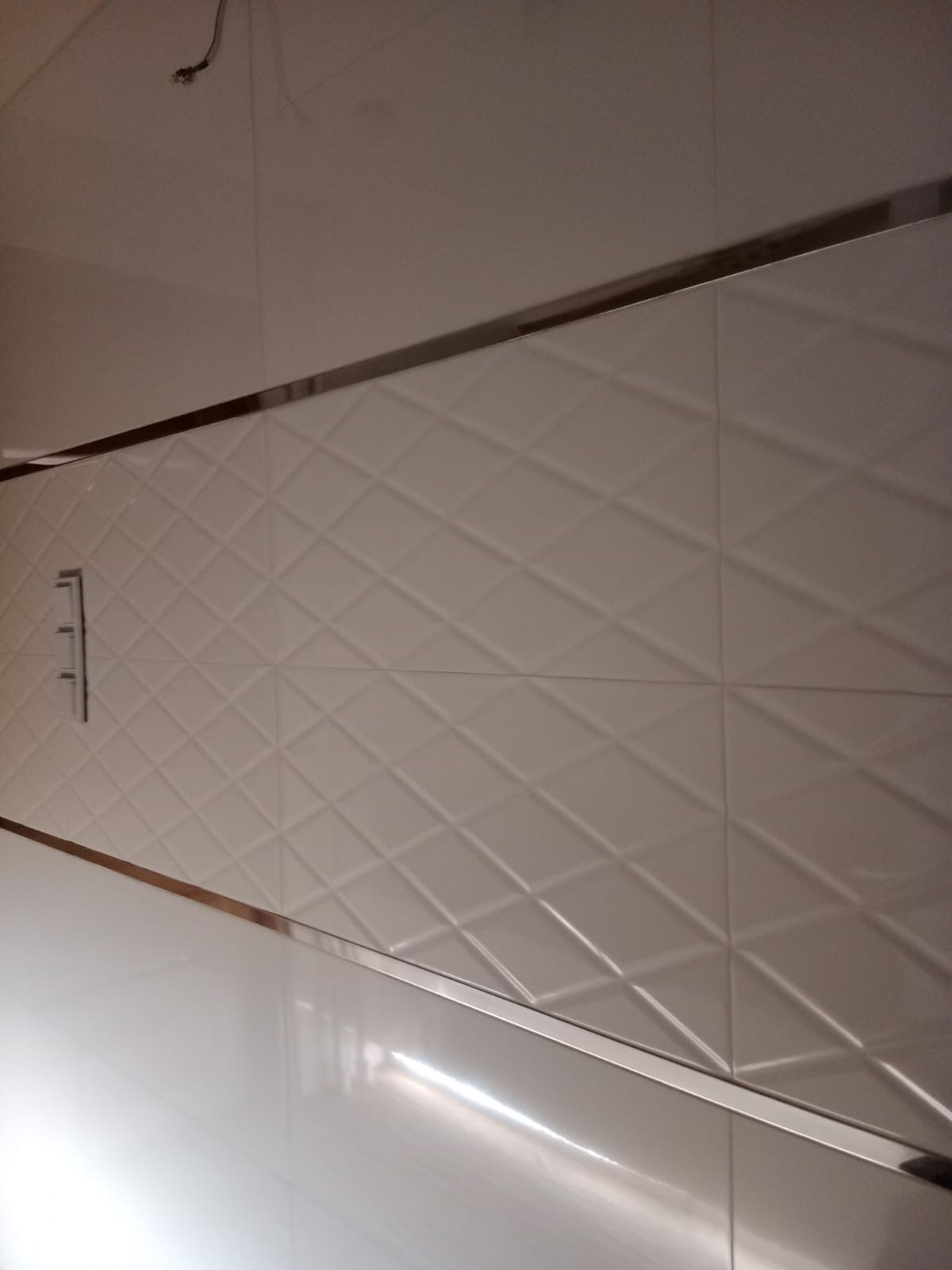 Białe, trójwymiarowe płytki ścienne ułożone w romby, oddzielone metalową listwą, z widocznym panelem sterowania.