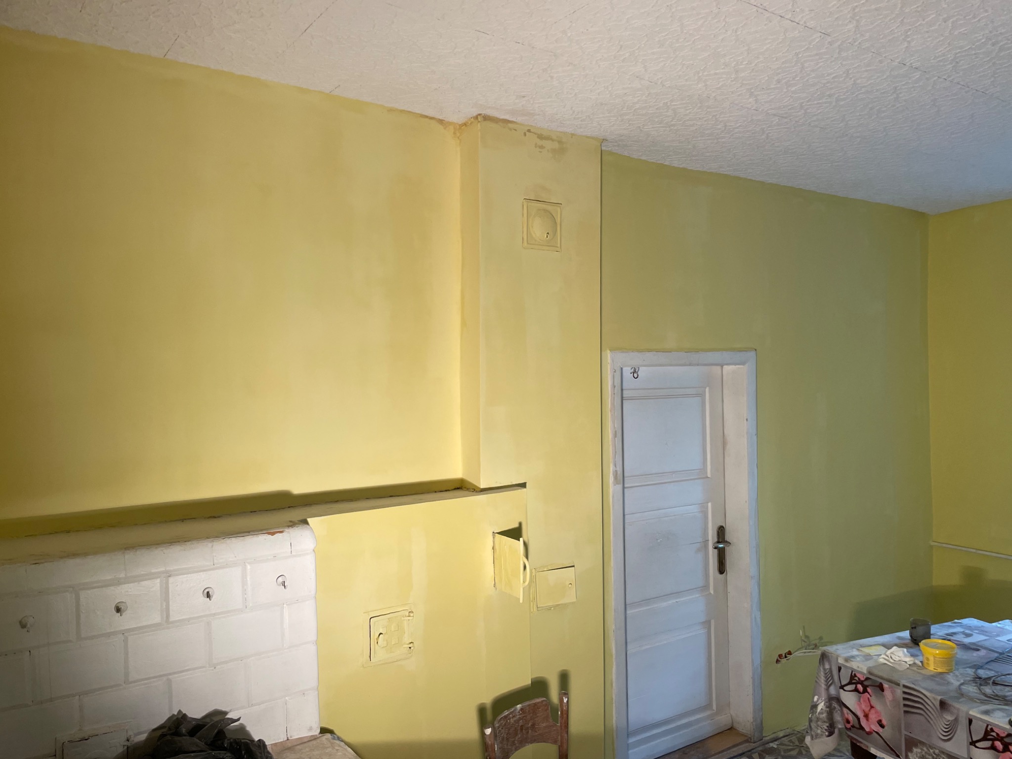 Żółte wnętrze pokoju po malowaniu, widoczny piec kaflowy z białymi kaflami, drewniane krzesło i stół z akcesoriami malarskimi.