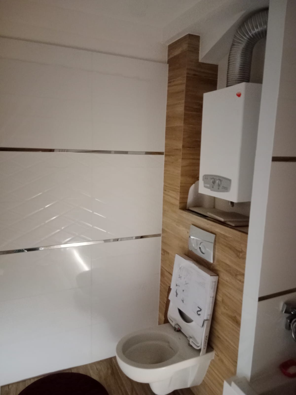 Świeżo wyremontowana łazienka z wiszącą toaletą i zabudowanym piecem gazowym, wykończona białymi płytkami z dekoracyjnymi paskami i panelami imitującymi drewno.