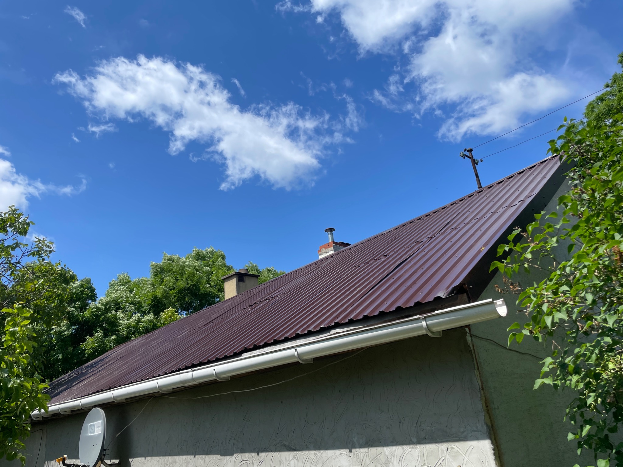 Brązowy dach z blachy trapezowej i białą rynną, widok z dołu na tle błękitnego nieba z obłokami, ściana budynku z anteną satelitarną po lewej stronie.