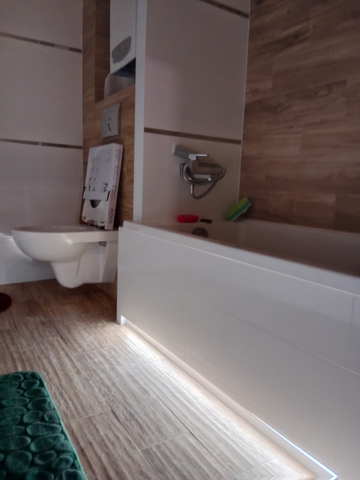 Nowoczesna łazienka z podwieszaną toaletą, białą wanną z podświetleniem LED u dołu i brązowymi płytkami imitującymi drewno na ścianach.