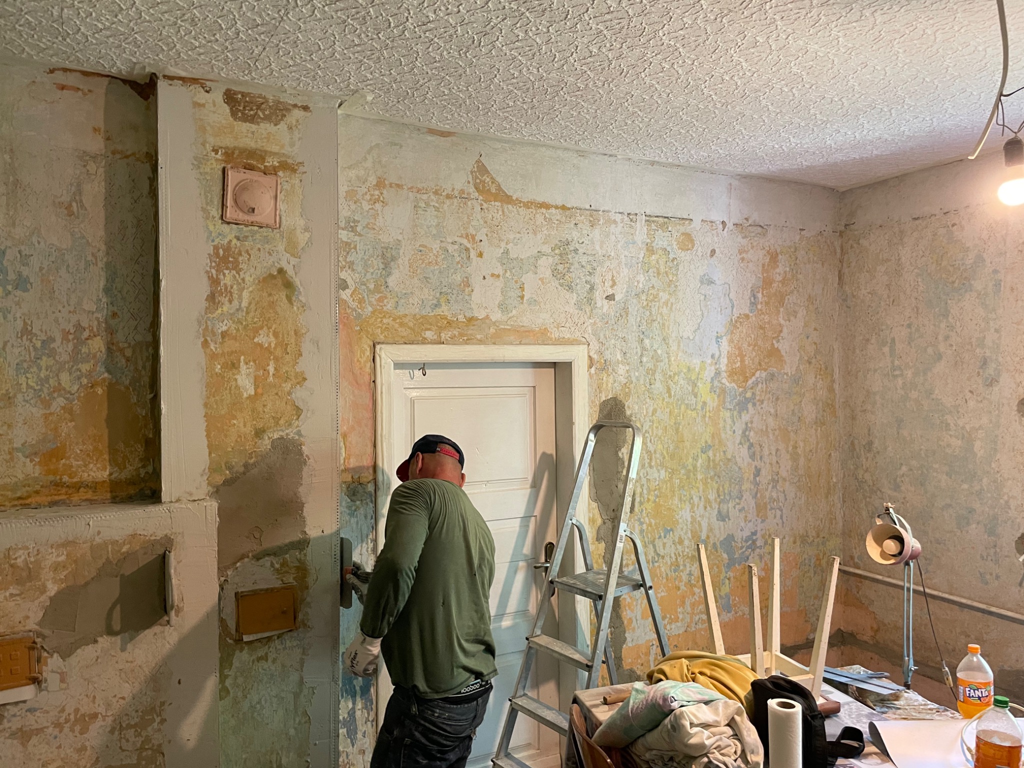 Mężczyzna w trakcie szpachlowania ściany w pomieszczeniu z widocznymi śladami starej farby i tapet, obok stoi drabina i stół z narzędziami oraz lampką.