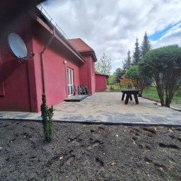 Mariusz Redmerski - Ułożona kostka brukowa w kolorze szarym tworząca taras przy czerwonym budynku. Widoczne drzewa i trawnik. Na tarasie stoi czarny stół.