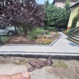 Mariusz Redmerski - Ścieżka z jasnej kostki z ciemną obwódką prowadząca przez ogród, widok z dołu. Fragment metalowej bramki i schody do domu w tle. Elementy ogrodu w tle.