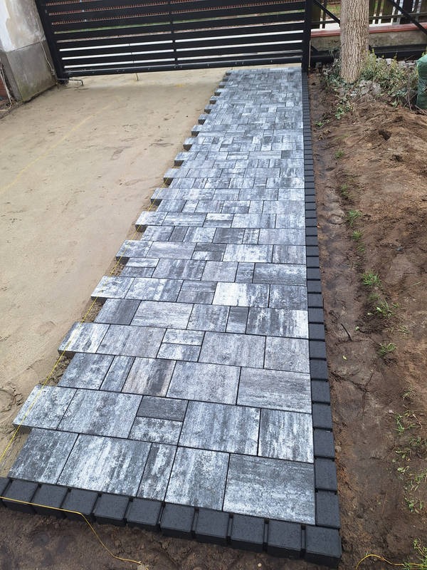 Ścieżka z szarej kostki brukowej o nieregularnym wzorze, obrzeża z ciemnej kostki, widoczna brama w tle, częściowo ułożona, żółta linka wyznaczająca linię.