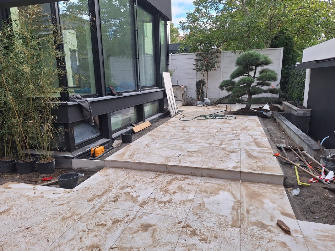 Jasne płytki tarasowe w trakcie układania, widoczne narzędzia i roślinność. Ujęcie z niskiej perspektywy, podkreślające detale powierzchni i otoczenia.