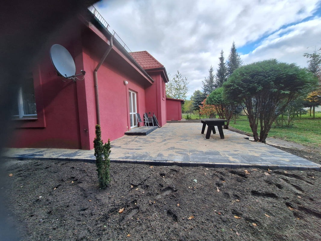 Ułożona kostka brukowa w kolorze szarym tworząca taras przy czerwonym budynku. Widoczne drzewa i trawnik. Na tarasie stoi czarny stół.