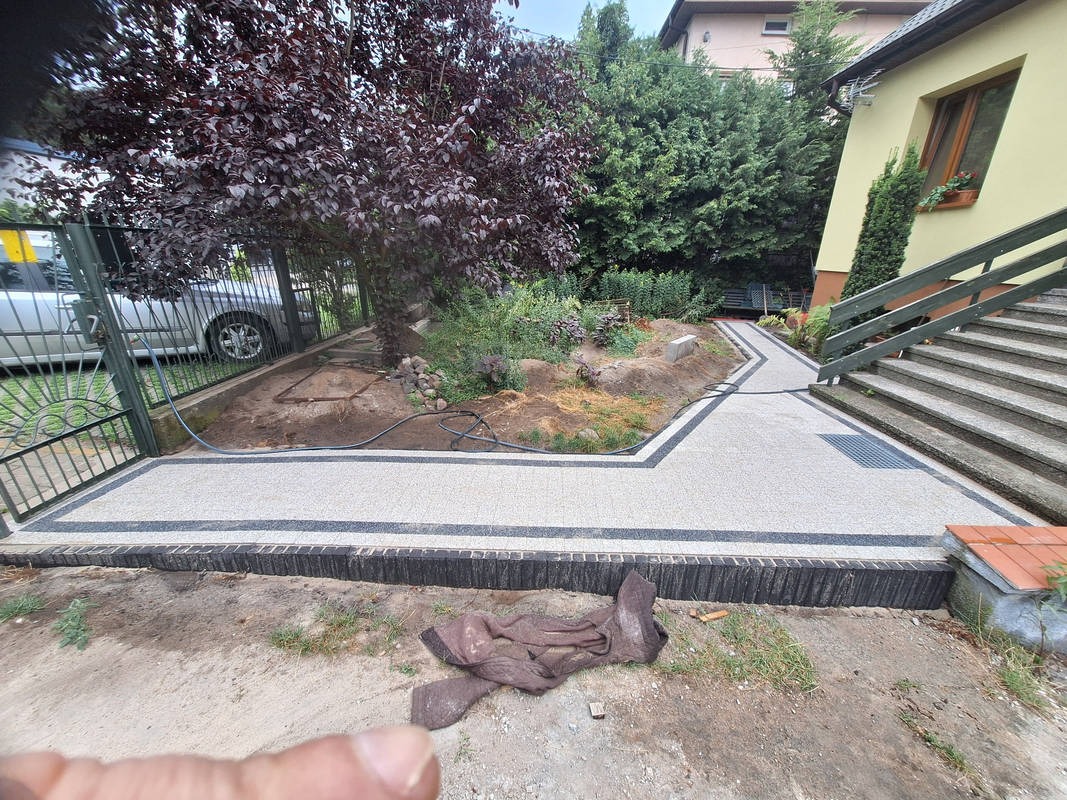 Ścieżka z jasnej kostki z ciemną obwódką prowadząca przez ogród, widok z dołu. Fragment metalowej bramki i schody do domu w tle. Elementy ogrodu w tle.