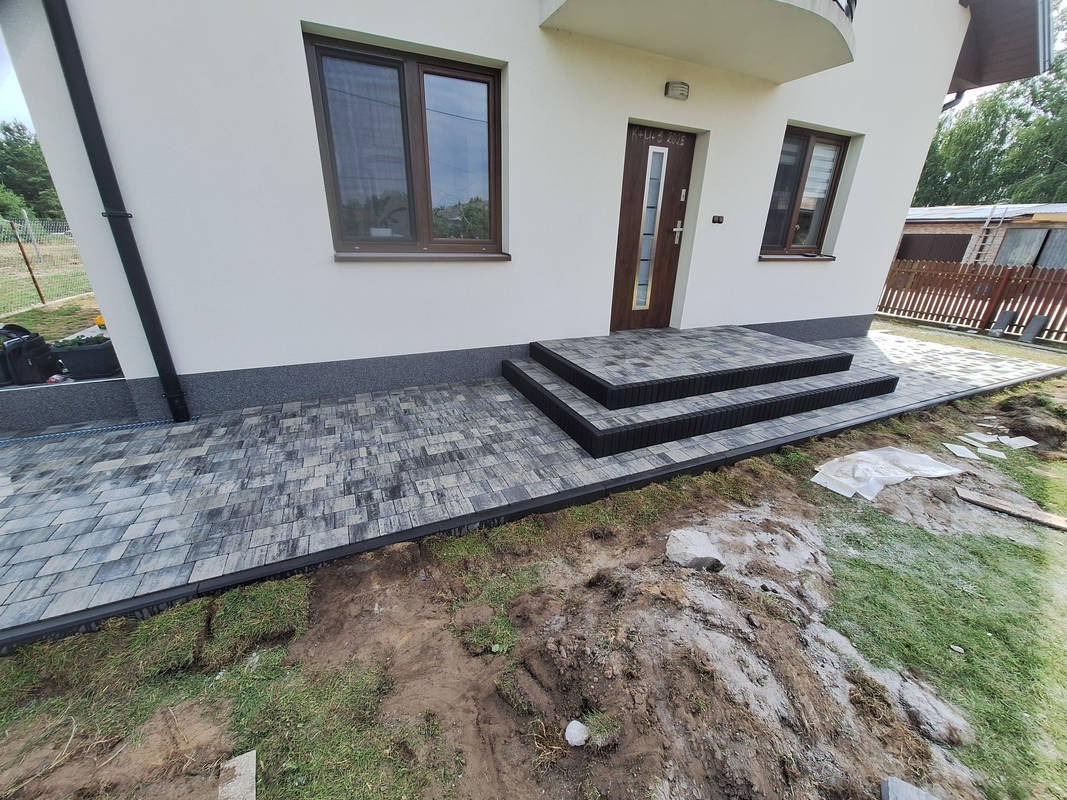 Ułożona kostka brukowa w szaro-czarnych odcieniach prowadząca do wejścia domu z trzema schodami. Widoczne fragmenty trawnika i ziemi.