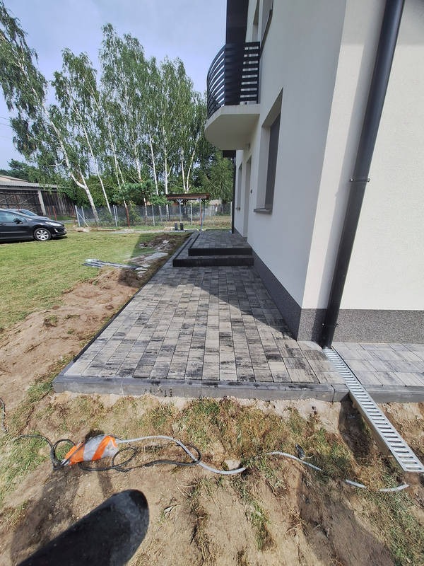 Ułożona kostka brukowa w szaro-białej kolorystyce prowadząca do schodów wejściowych domu z widocznym odpływem liniowym i kablem elektrycznym na ziemi.