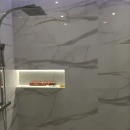 Nowoczesna łazienka z kabiną prysznicową typu deszczownica, wykończona białymi płytkami imitującymi marmur, z podświetloną wnęką, w której leży poziomica i taśma malarska.