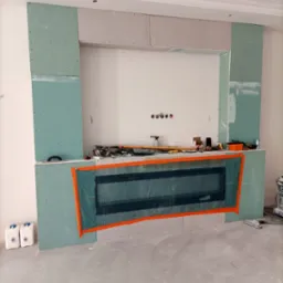 Ściana z zamontowanym wkładem kominkowym, obudowana płytami kartonowo-gipsowymi w trakcie montażu, widoczne narzędzia i materiały budowlane, zabezpieczony wkład kominkowy folią ochronną.