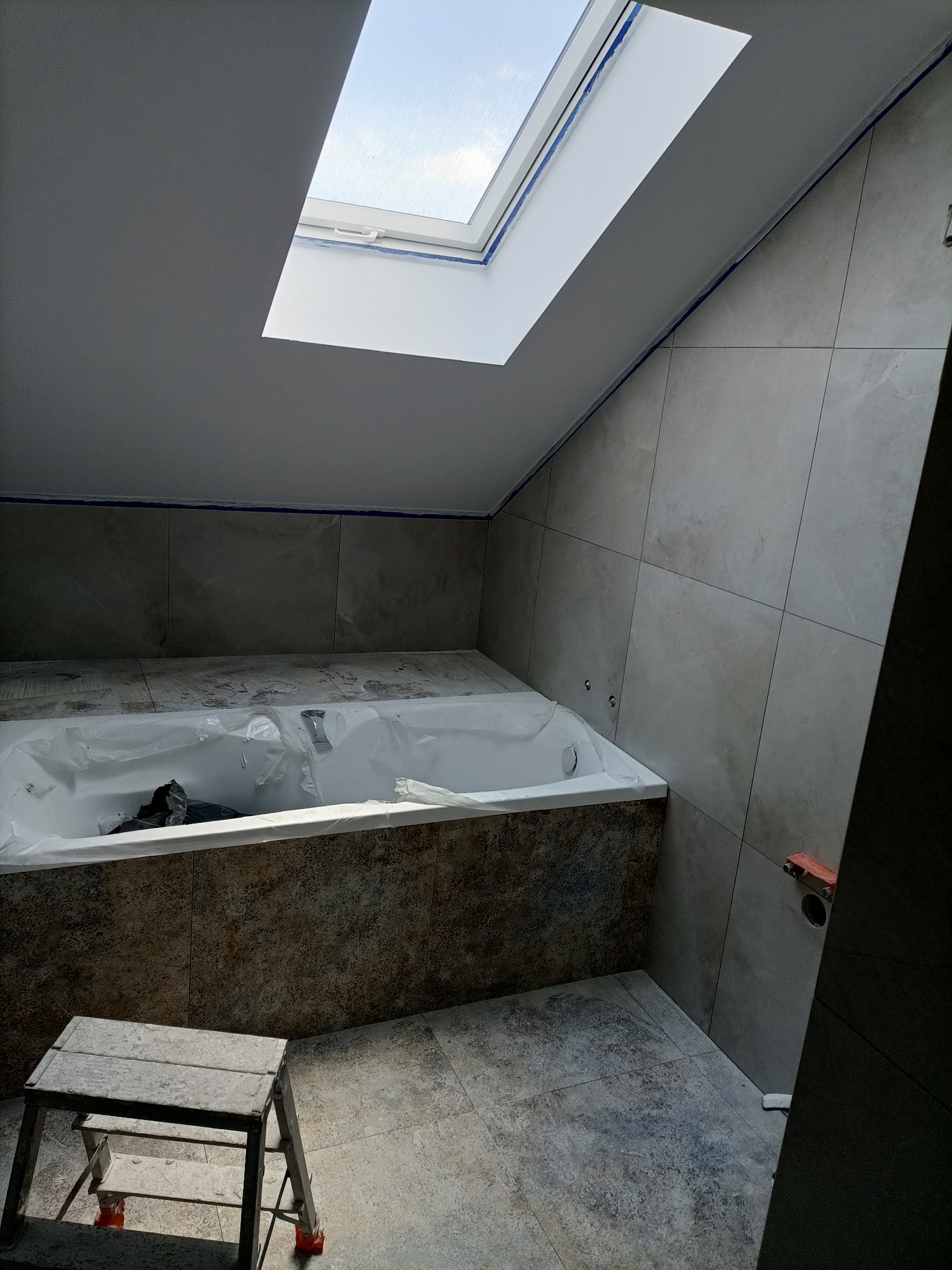 Łazienka na poddaszu w trakcie remontu: wanna zabezpieczona folią, ułożone płytki ścienne i podłogowe w odcieniach szarości, widoczna drabinka i okno dachowe z niebieską taśmą malarską.