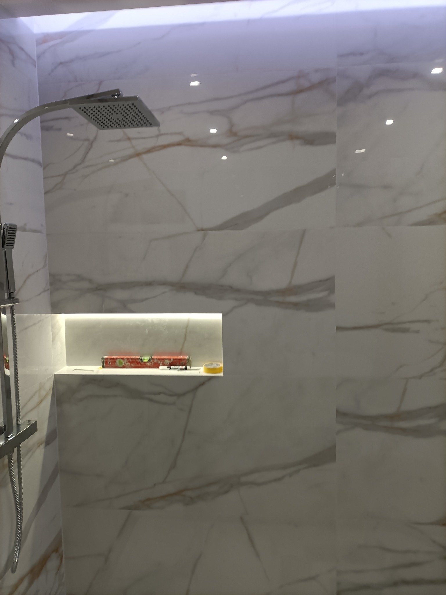 Nowoczesna łazienka z kabiną prysznicową typu deszczownica, wykończona białymi płytkami imitującymi marmur, z podświetloną wnęką, w której leży poziomica i taśma malarska.