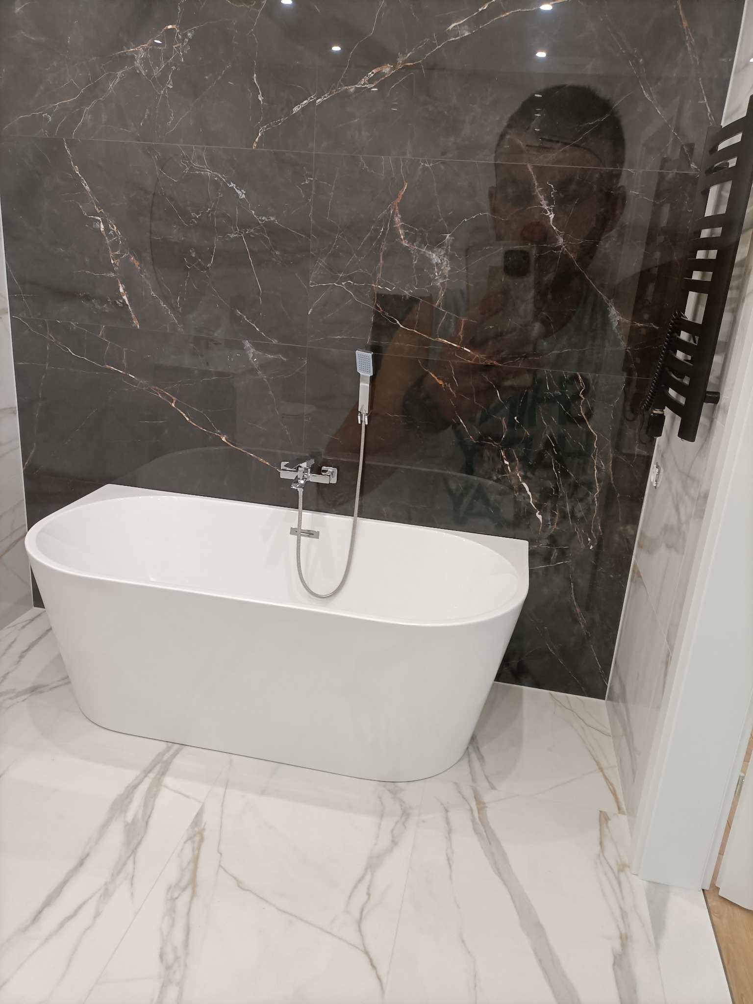 Nowoczesna łazienka z wolnostojącą białą wanną na tle ściany z ciemnego marmuru z złotymi żyłami, widoczne odbicie osoby robiącej zdjęcie w lustrzanej powierzchni.