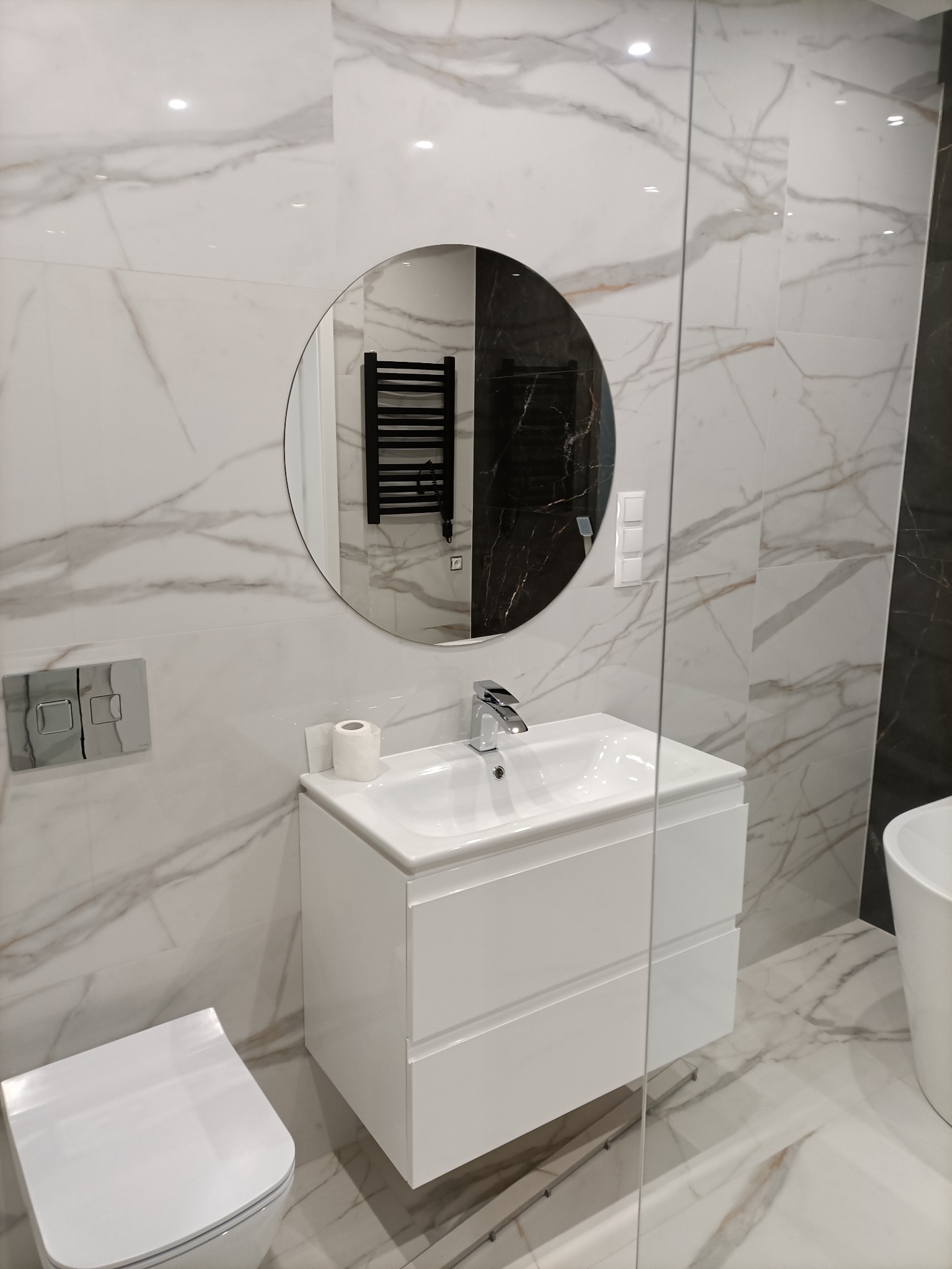 Nowoczesna łazienka z białymi marmurkowymi płytkami, okrągłym lustrem odbijającym czarny grzejnik drabinkowy, białą szafką pod umywalką i wiszącą toaletą.
