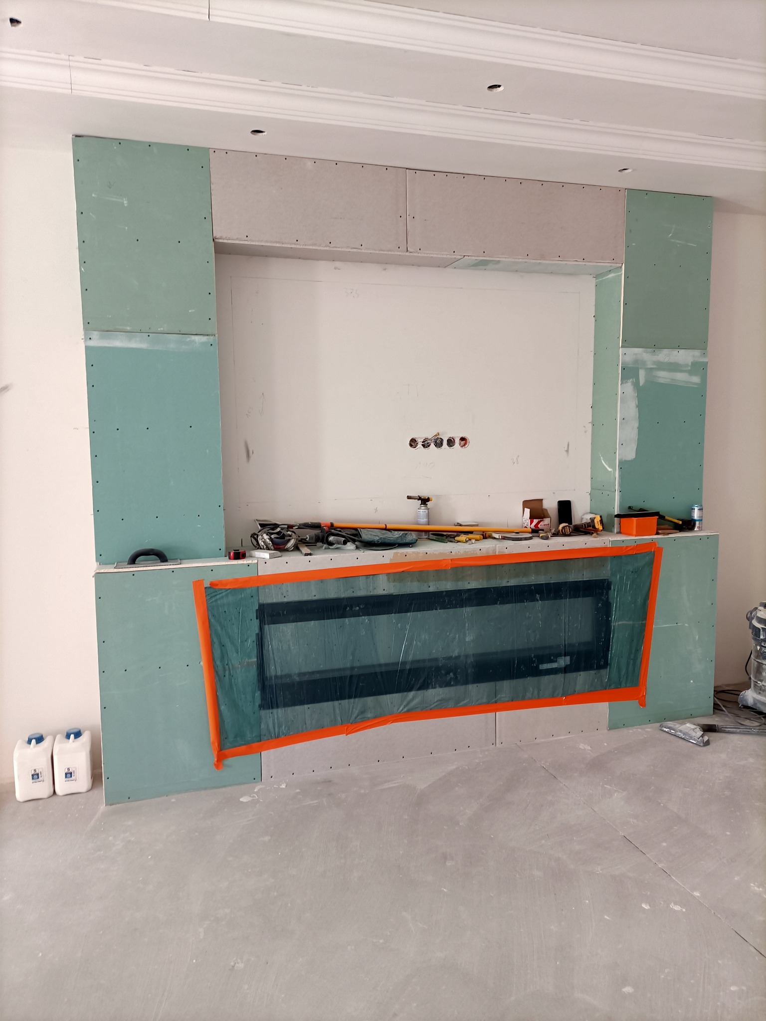 Ściana z zamontowanym wkładem kominkowym, obudowana płytami kartonowo-gipsowymi w trakcie montażu, widoczne narzędzia i materiały budowlane, zabezpieczony wkład kominkowy folią ochronną.