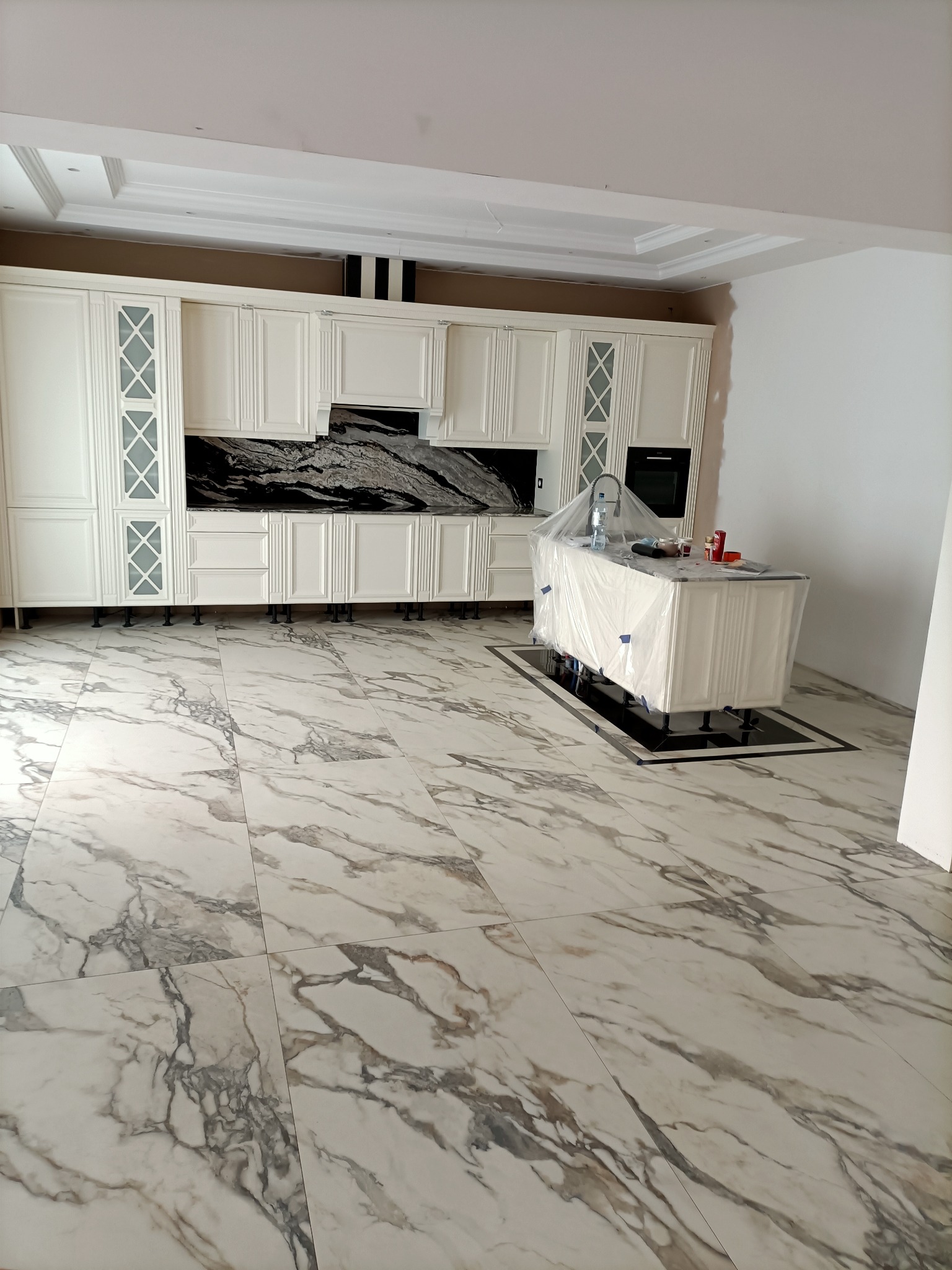 Wnętrze kuchni w trakcie montażu: białe szafki z przeszklonymi drzwiczkami, czarny blat z marmurowym wzorem, wyspa kuchenna przykryta folią ochronną, podłoga wyłożona białymi płytkami z szarymi...