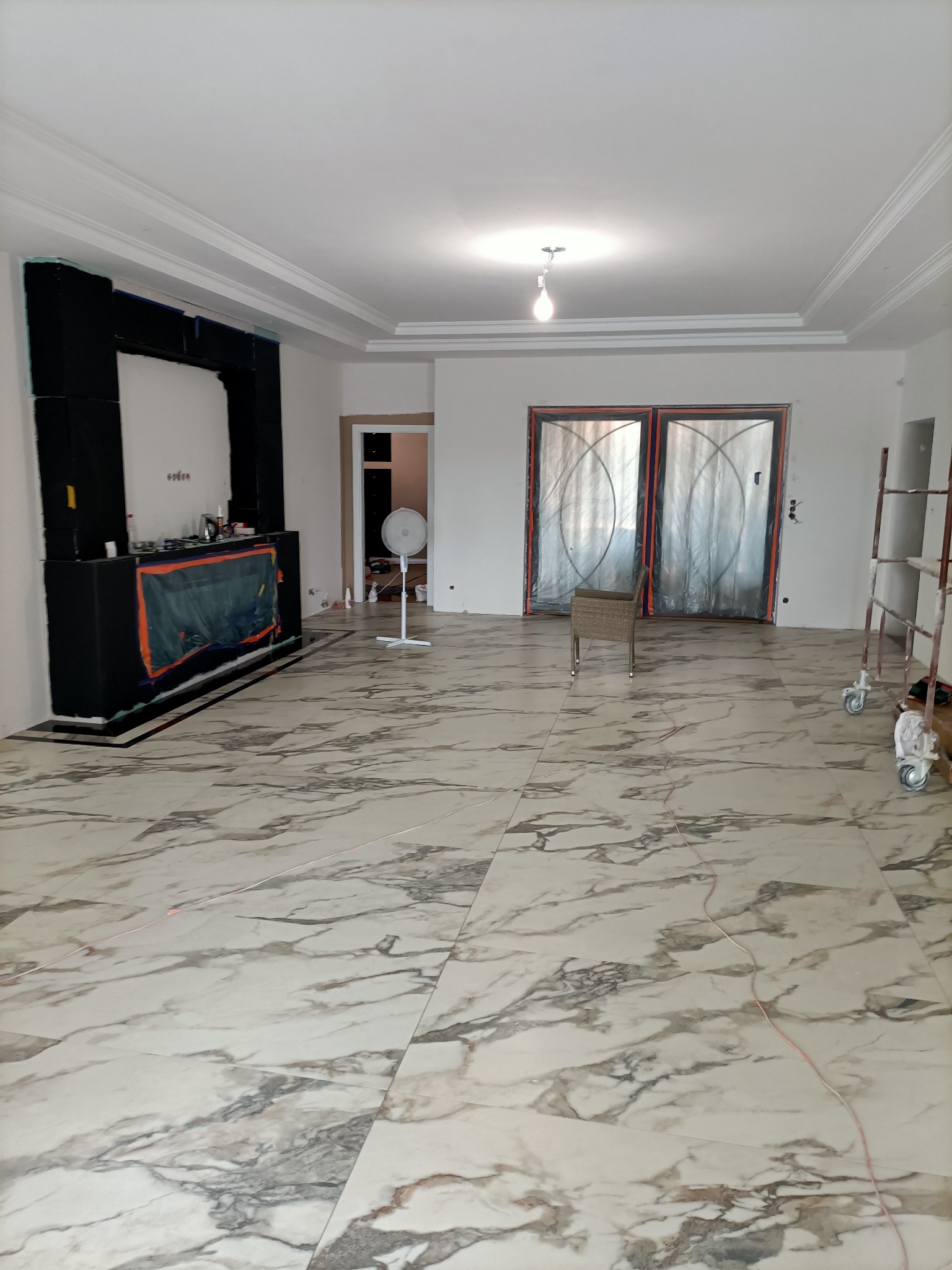 Wnętrze w trakcie remontu: marmurowe płytki podłogowe z laserowo wyznaczonymi liniami, osłonięte drzwi balkonowe, zabezpieczona zabudowa ściany, wentylator stojący i drabina na kółkach.