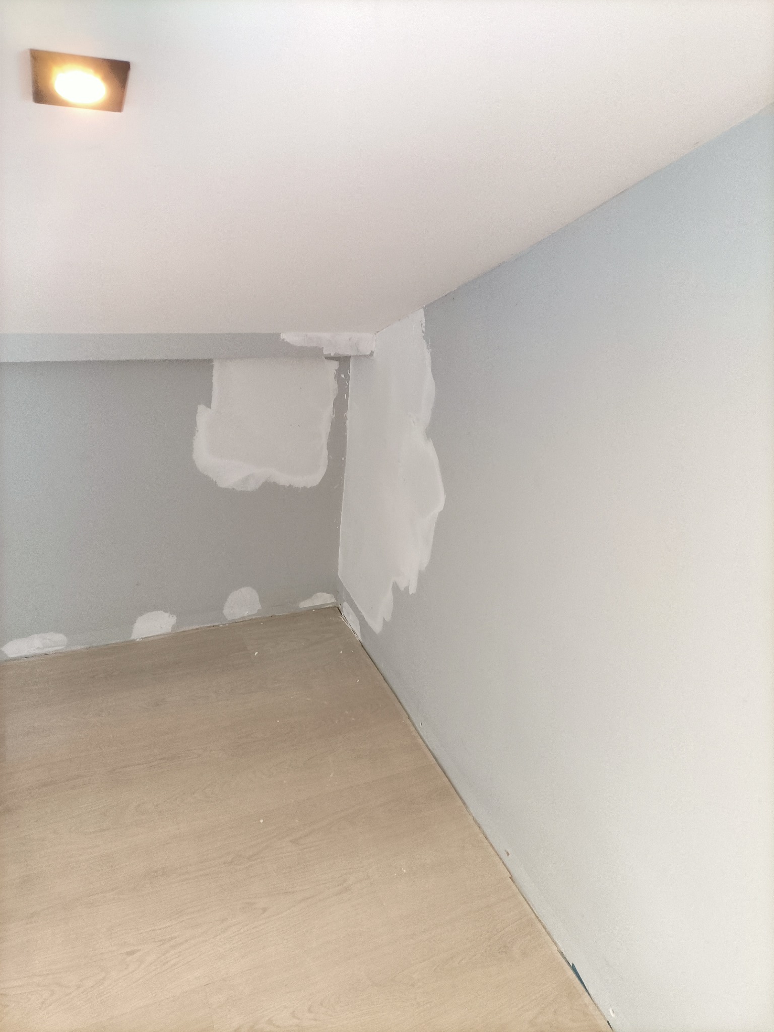 Pokój w trakcie remontu, ściany z szarym tłem częściowo pokryte gładzią, widoczne fragmenty podłogi z panelami i lampa sufitowa.