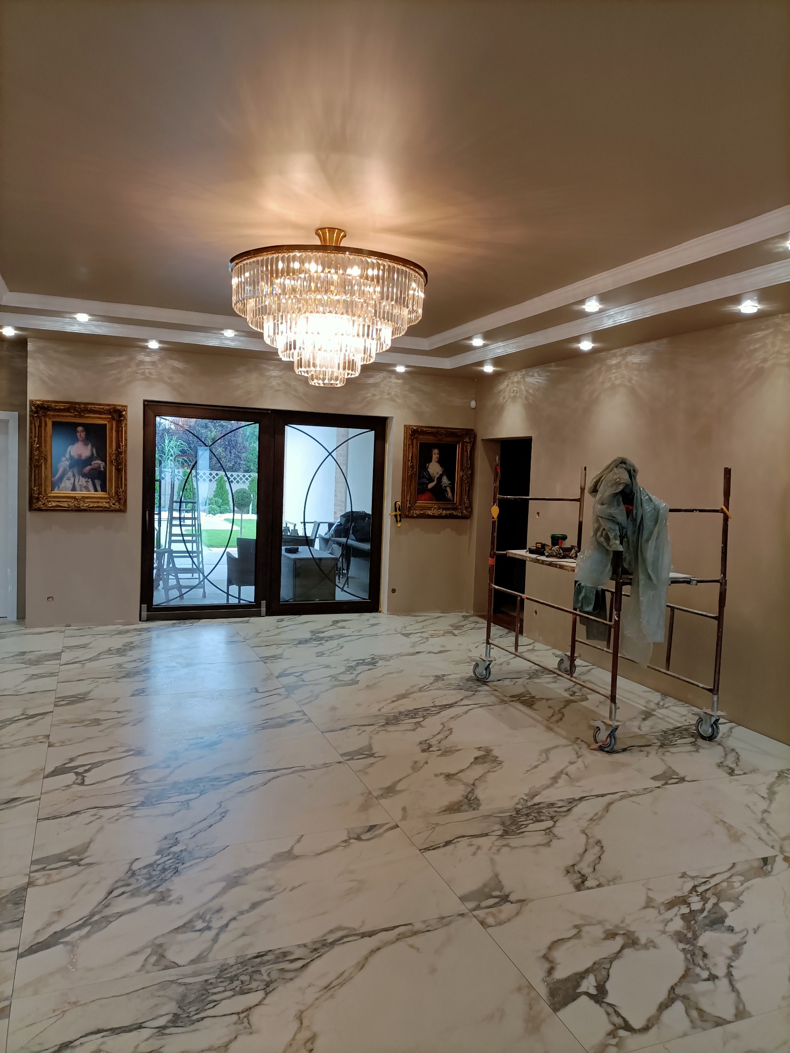 Wnętrze pokoju z marmurową podłogą, klasycznym żyrandolem i sztukaterią, ściany w beżowym odcieniu, widoczne rusztowanie z narzędziami sugerujące prace wykończeniowe.