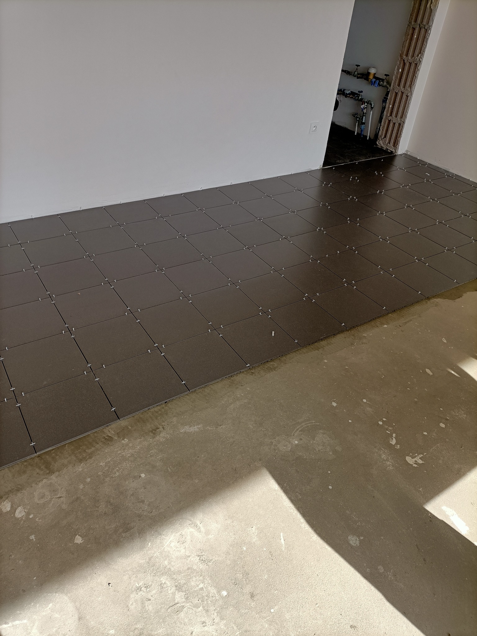 Proces układania ciemnobrązowych płytek podłogowych w karo, z widocznymi krzyżykami dystansowymi, na częściowo przygotowanym podłożu betonowym w jasnym pomieszczeniu z instalacją hydrauliczną w tle.