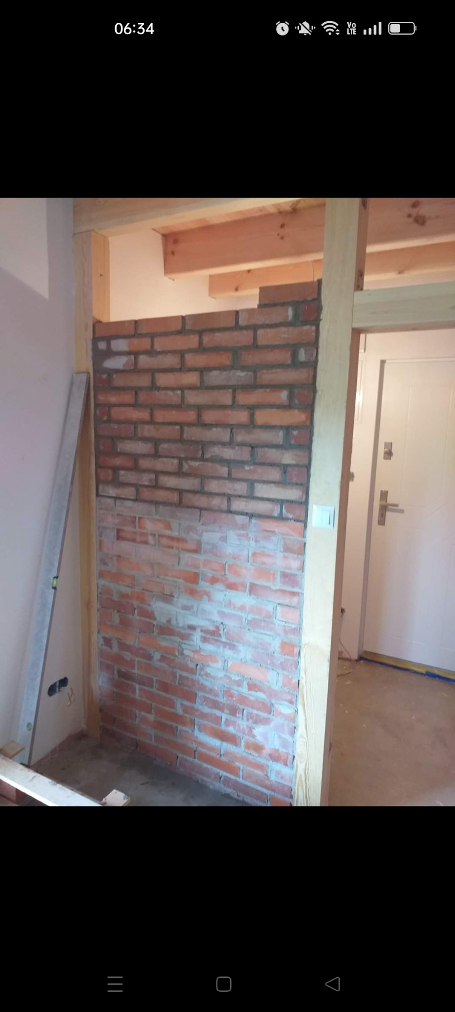 Częściowo wykończona ściana z czerwonej cegły wewnątrz pomieszczenia, widoczne drewniane elementy konstrukcyjne, poziomica i fragment białych drzwi.