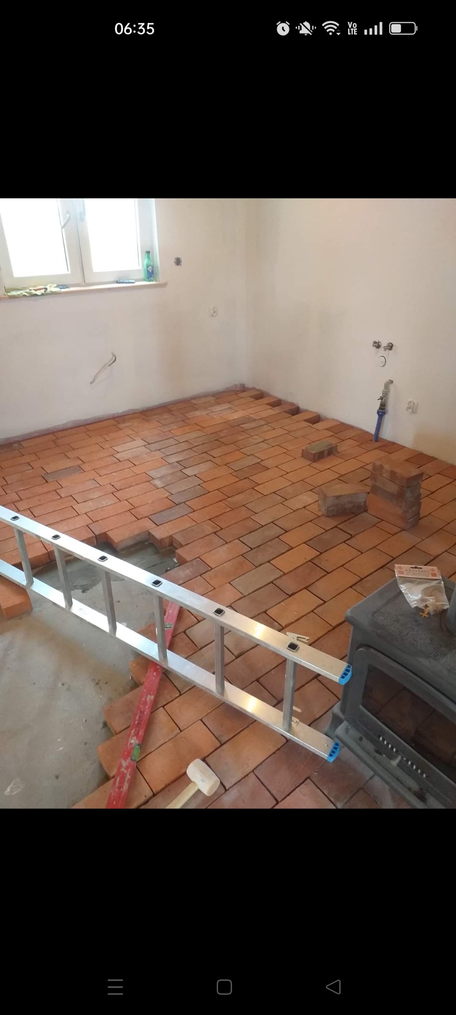 Układanie ceglastej terakoty na podłodze w trakcie remontu, widoczna drabina aluminiowa, poziomica i guma młotek, w tle piec wolnostojący.