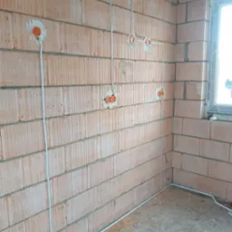 Surowe wnętrze w Gryficach z widoczną instalacją elektryczną w peszlach, zamontowaną na ścianie z cegły dziurawki przed tynkowaniem, z widocznymi puszkami elektrycznymi.