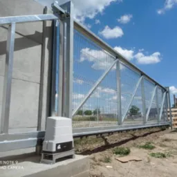 Przesuwna brama z metalowej siatki, z automatyką napędu Nice, zamontowana na betonowym fundamencie, w tle błękitne niebo z chmurami.