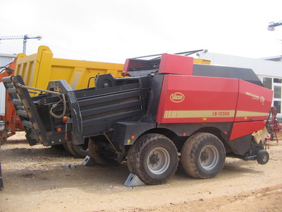 Czerwono-czarna prasa rolująca Vicon LB 12200 stoi na ziemi, zabezpieczona klinami pod kołami, z żółtą wywrotką w tle.