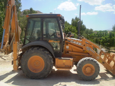 Pomarańczowa koparko-ładowarka CASE 580 Super R Series 2, zaparkowana na placu z widocznym drzewem w tle.