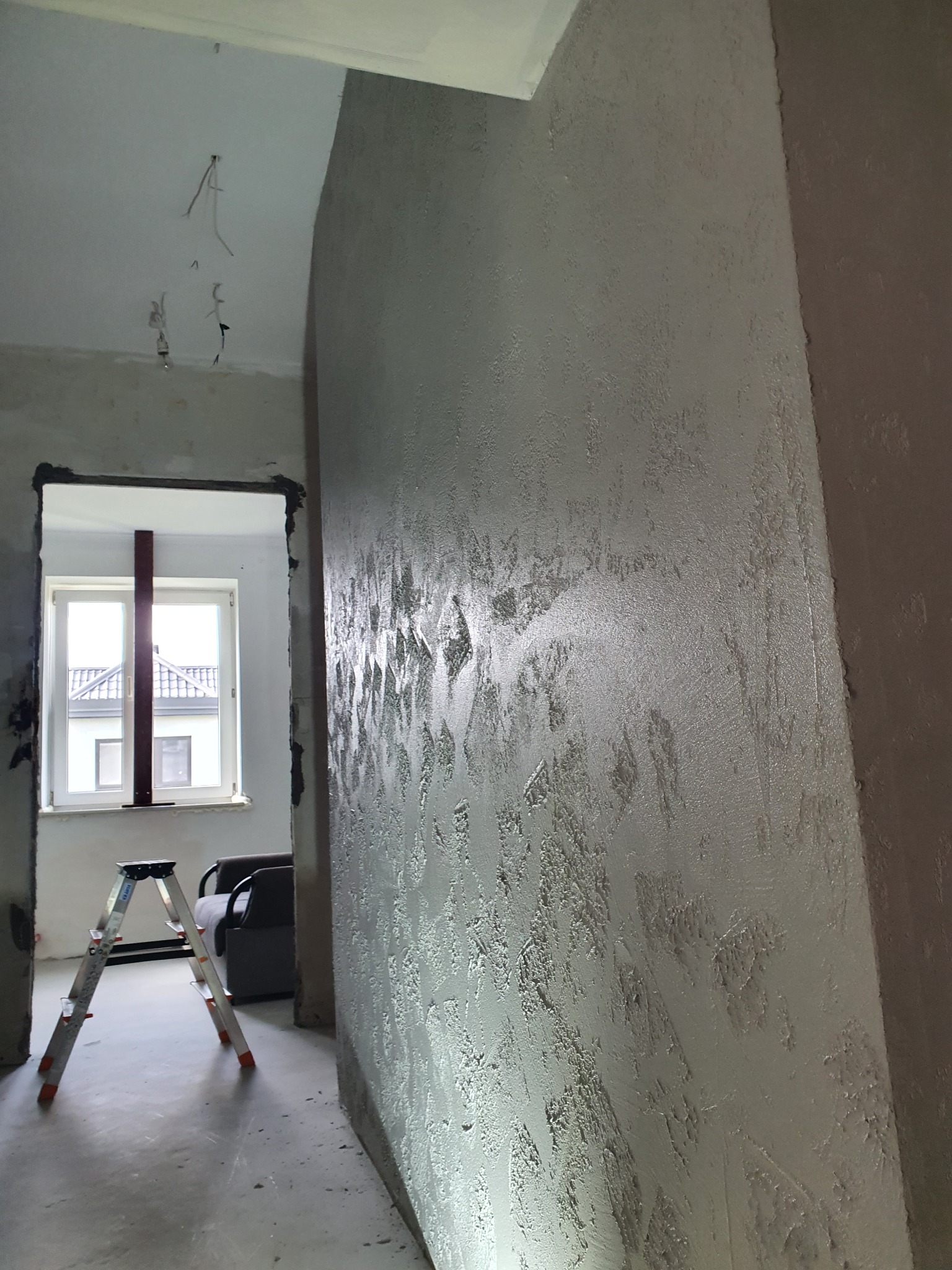 Ściana z efektem betonu architektonicznego w trakcie wykańczania, widoczny fragment drabiny malarskiej i otwór drzwiowy z przewodami wystającymi ze ściany.