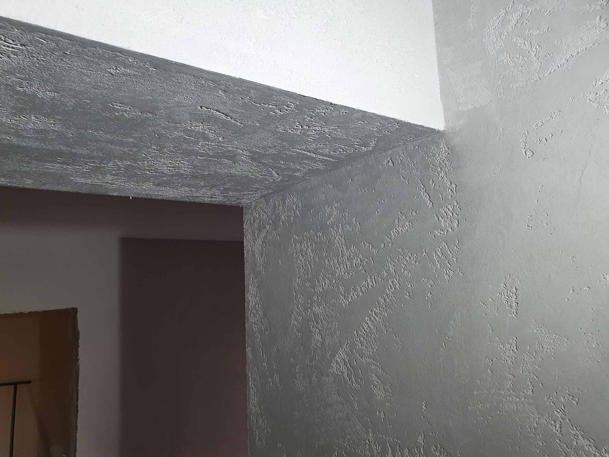 Szara ściana i sufit pokryte dekoracyjnym betonem architektonicznym z wyraźną fakturą, widoczne z bliska w rogu pomieszczenia.