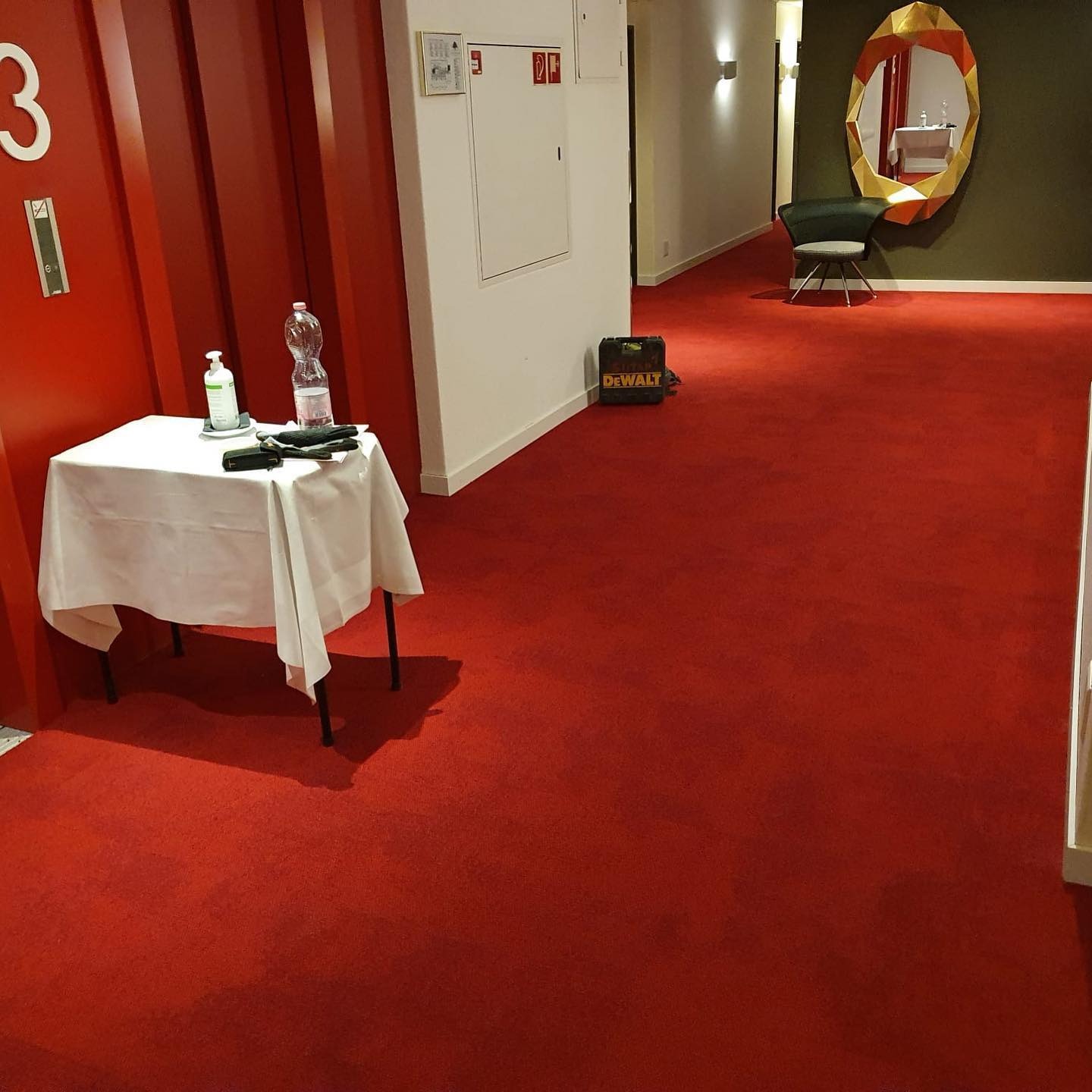 Czerwona wykładzina dywanowa w korytarzu hotelowym, widoczny stolik z płynem do dezynfekcji i butelką wody, w tle skrzynka narzędziowa DeWalt.