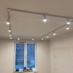 Przedsiębiorca Lidia Geers Remonty i Wykończenia - Białe wnętrze z zamontowanym systemem szynowym z reflektorami punktowymi na suficie, widok na okno z narzędziami na parapecie i dekoracyjną ścianą z pionowymi żłobieniami.