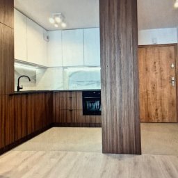 Przedsiębiorca Lidia Geers Remonty i Wykończenia - Nowoczesna kuchnia z drewnianymi szafkami, białymi górnymi szafkami i czarną armaturą, widok na drewniane drzwi wejściowe.