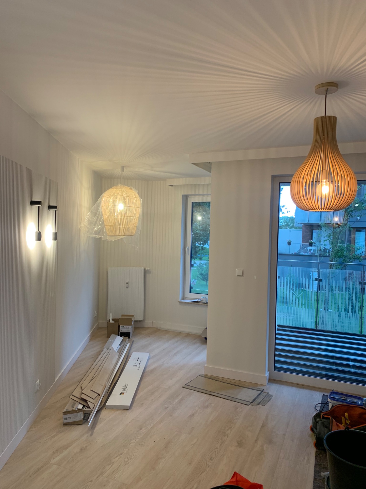 Wnętrze pokoju z drewnianą podłogą w trakcie prac wykończeniowych, widoczne panele podłogowe, wiszące lampy z drewnianymi abażurami i nowoczesne oświetlenie ścienne.