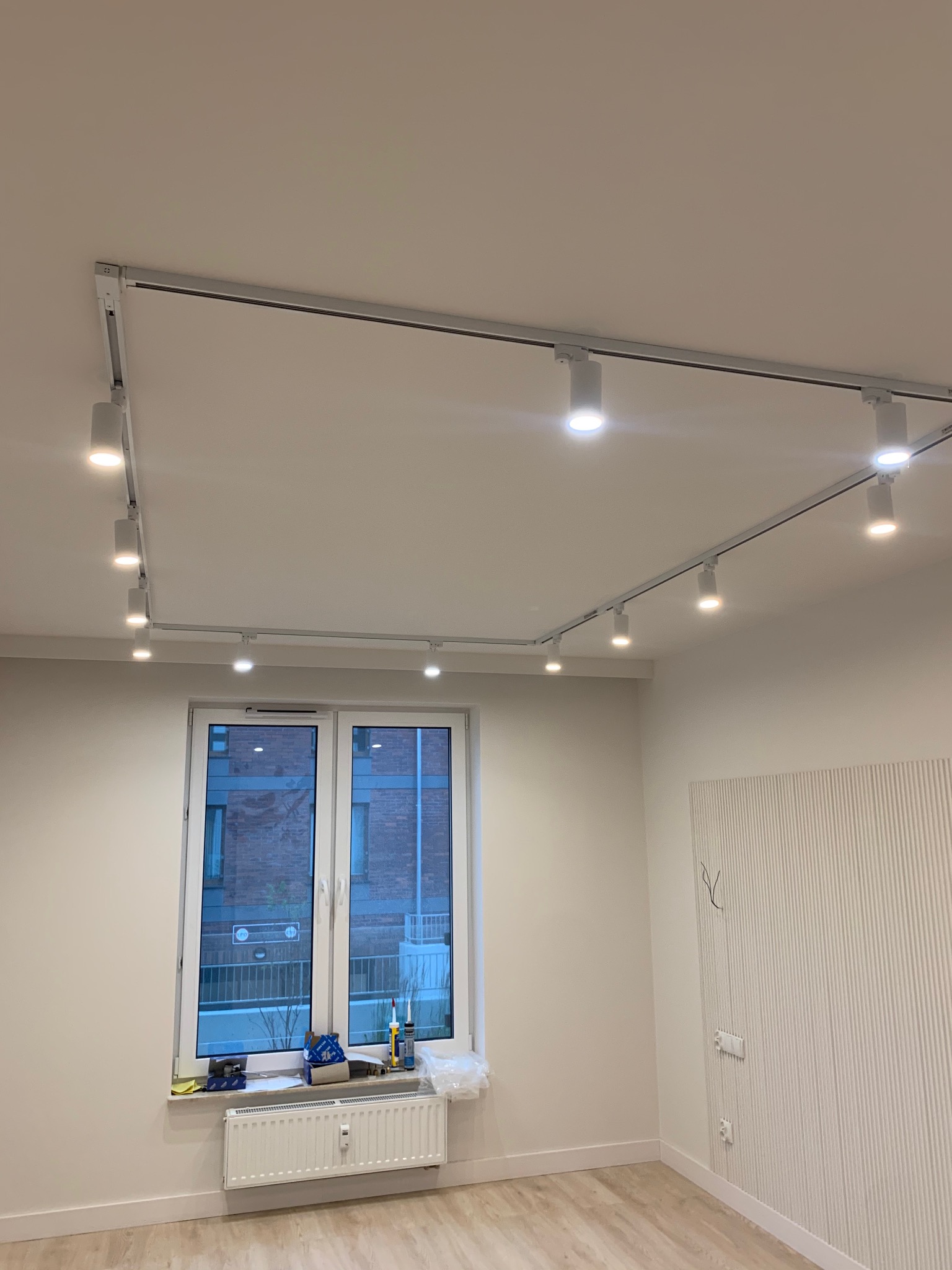 Białe wnętrze z zamontowanym systemem szynowym z reflektorami punktowymi na suficie, widok na okno z narzędziami na parapecie i dekoracyjną ścianą z pionowymi żłobieniami.