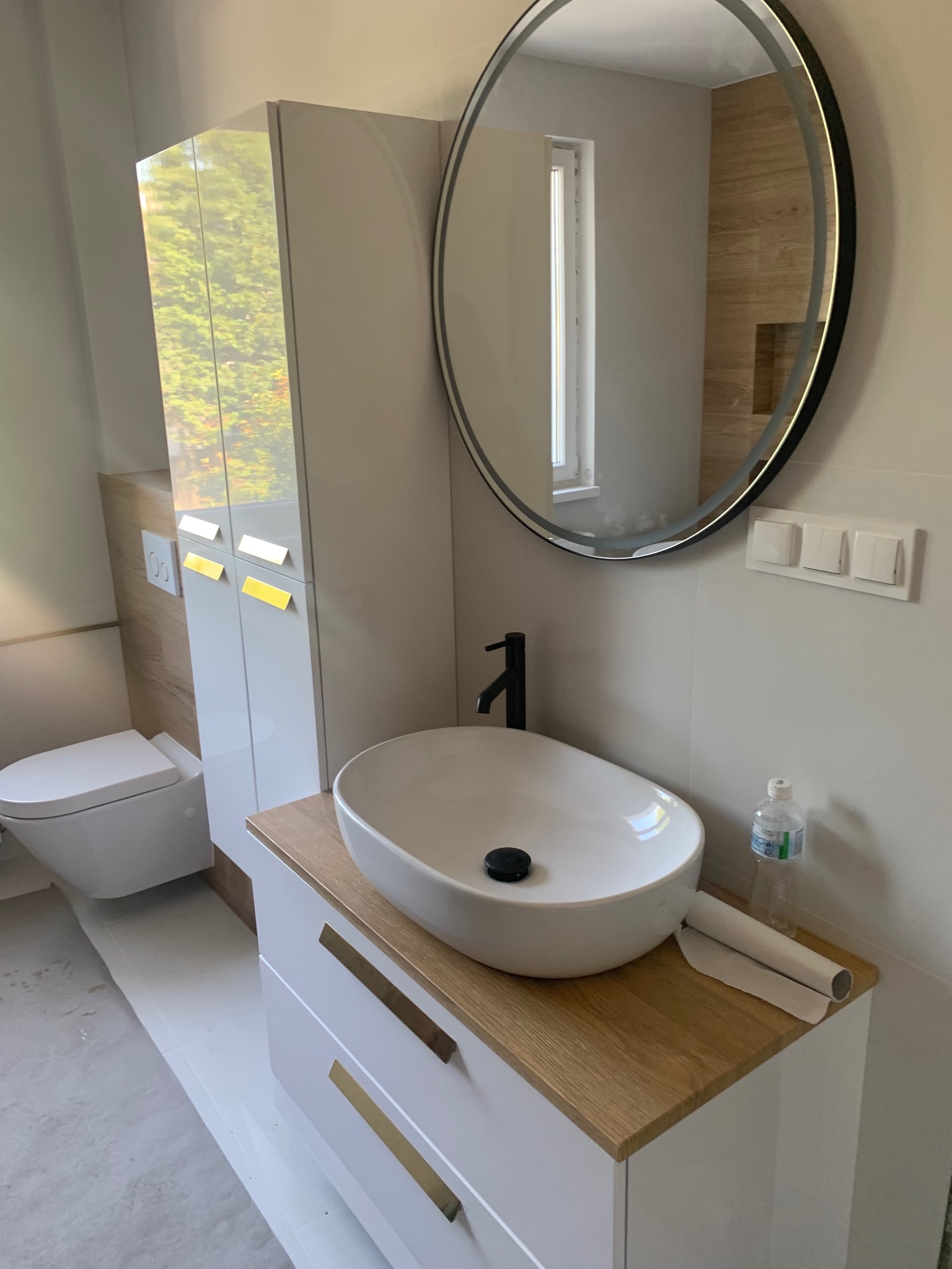 Nowoczesna łazienka w Niekłonicach z okrągłym lustrem z podświetleniem LED, białą umywalką nablatową na szafce z drewnianym blatem i złotymi uchwytami, oraz wiszącą toaletą.